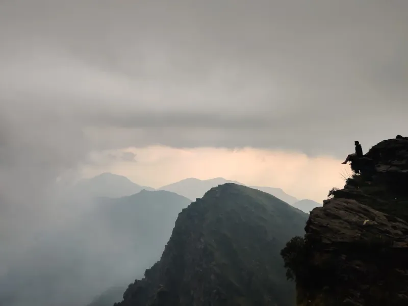 A trekker soaking in dramatic cloud views from Chandrashila summit on the Chopta trek. | © MountRoutes