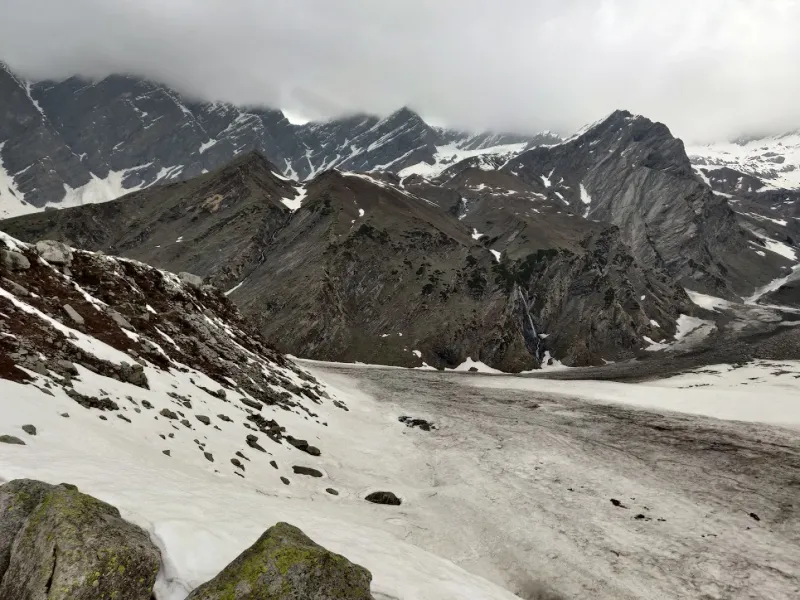 Nature's scale — a distant mountain waterfall dwarfed by the towering snowfields of the Manali range. | © MountRoutes